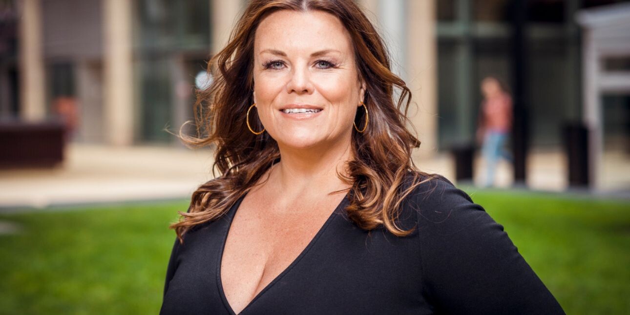 A portrait of Natalie Rogers. Natalie is a white woman with long brown hair wearing a v-necked black top. There is a lawn and buildings in the background
